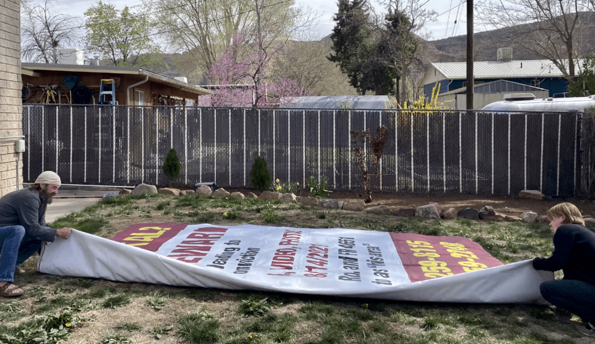 Two people are unfurling a massive sign; the sign takes up most of the backyard they are in.