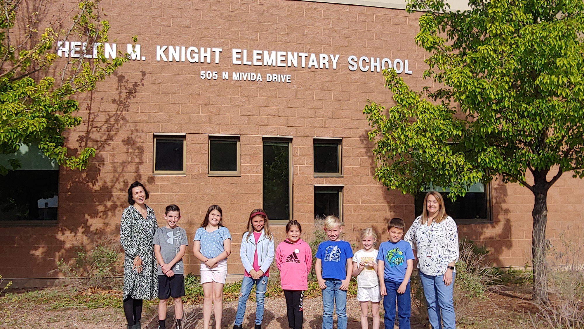 The principals stand on either end of a line of children in front of HMK Elementary. They are all squinting due to facing the sun, but are smiling!