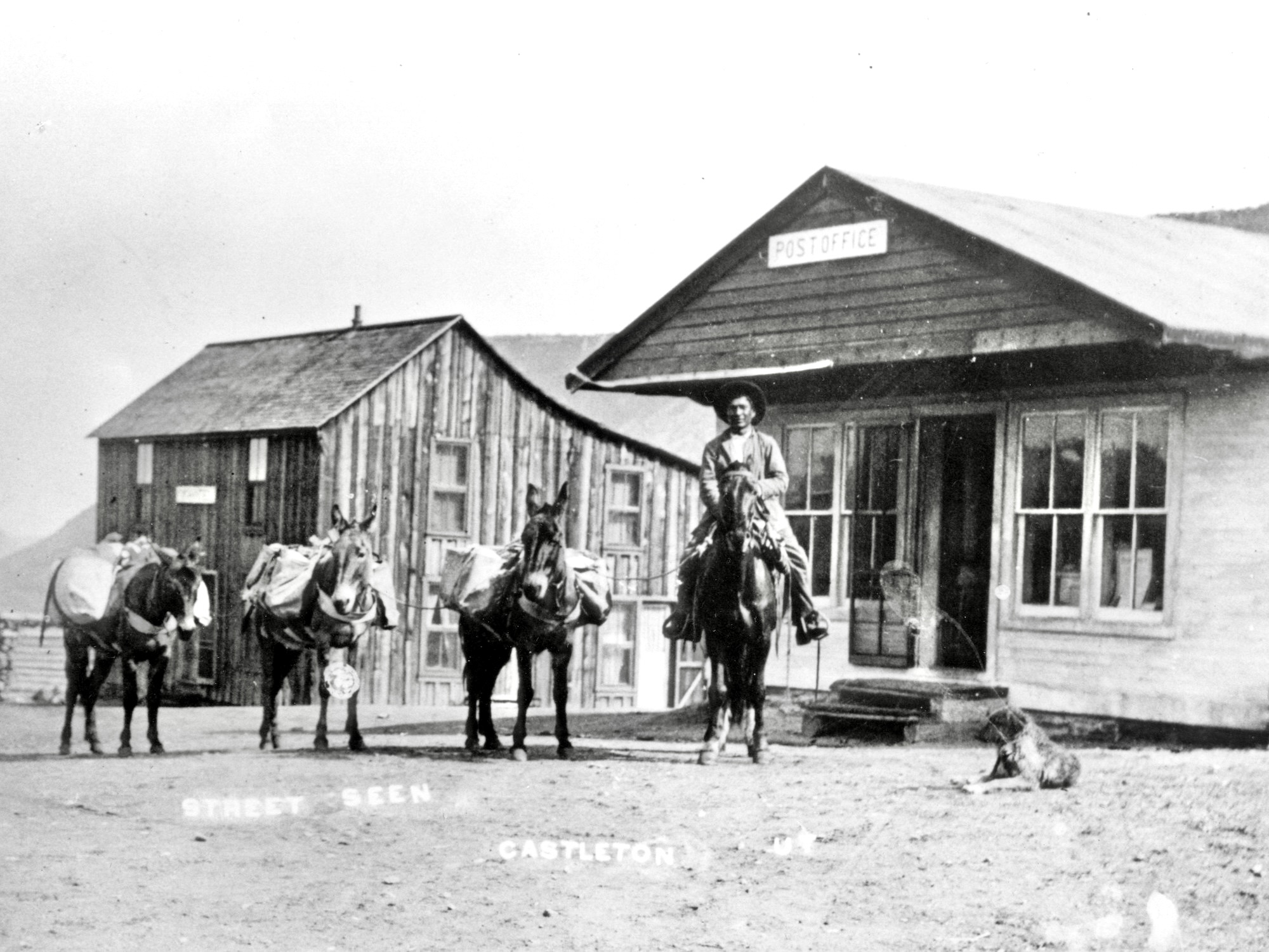 A cowboy on his horse in front of the post office, with three donkeys. The mules have packs. The post office building behind them is wooden and small, with four windows on its front and an open door. There is a small sign on the building that says "Post Office"