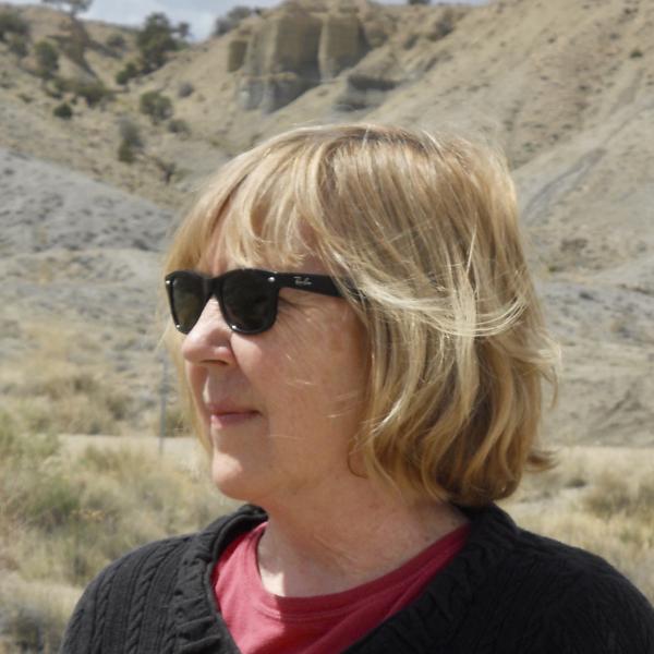 Nancy Takacs has a shoulder-length blonde bob; here, she's wearing sunglasses and standing amongst sand dunes.