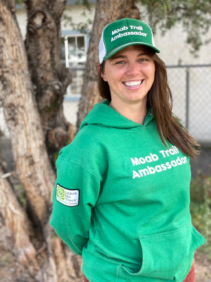 Anna Sprout has a huge grin and is wearing her trail ambassador outfit: a green hat and hoodie that say "Moab Trail Ambassador"