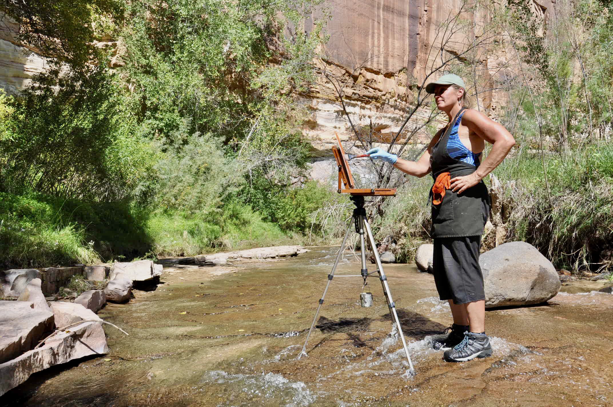 Loretta Domaszewski stands in a river in her Keens with an easel and apron. She's looking up at the canyon walls surrounding her and painting.