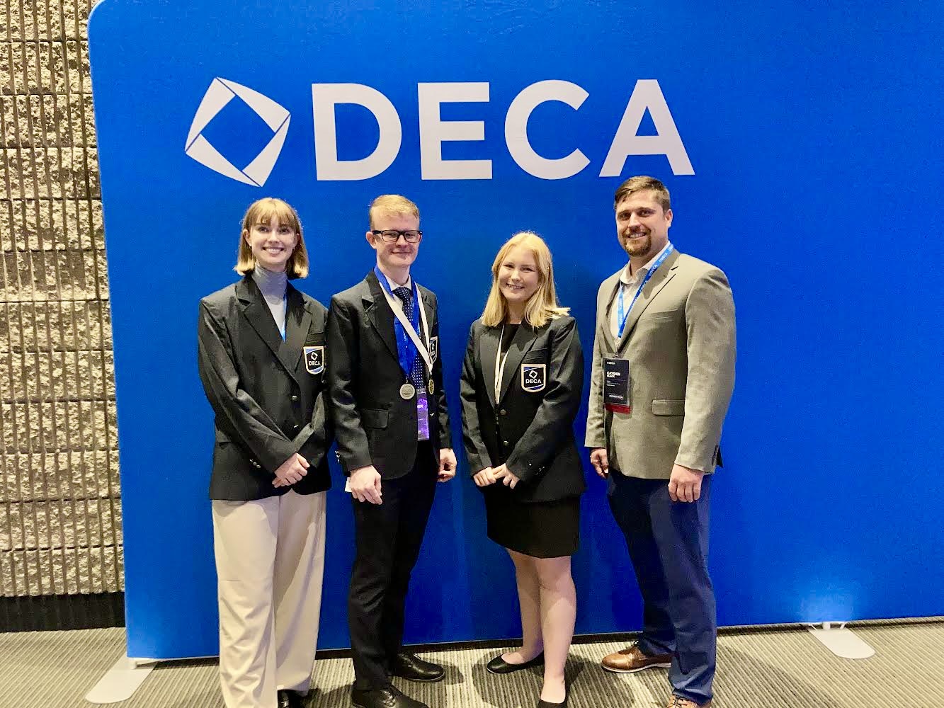 Lillian, Alex, Kaistin, and their advisor Cayden Black pose in front of a large blue wall that says "DECA."
