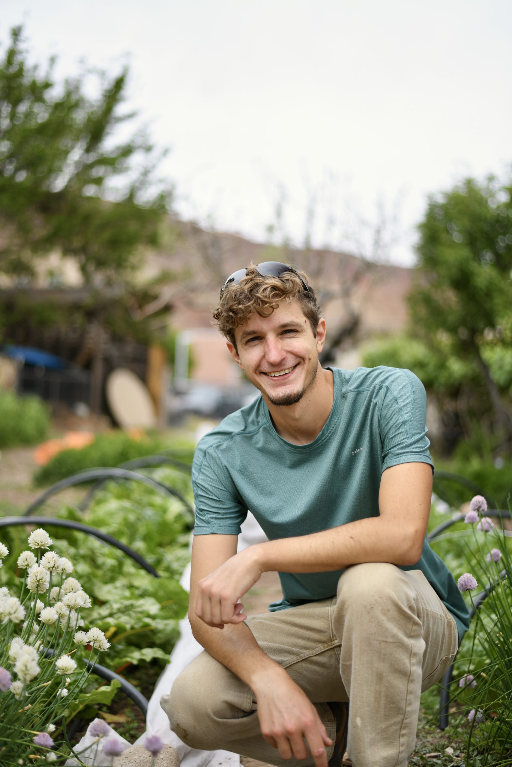 Ben Menard is crouching down among the garden rows at the Youth Garden Project. He has curly hair and a soft smile.