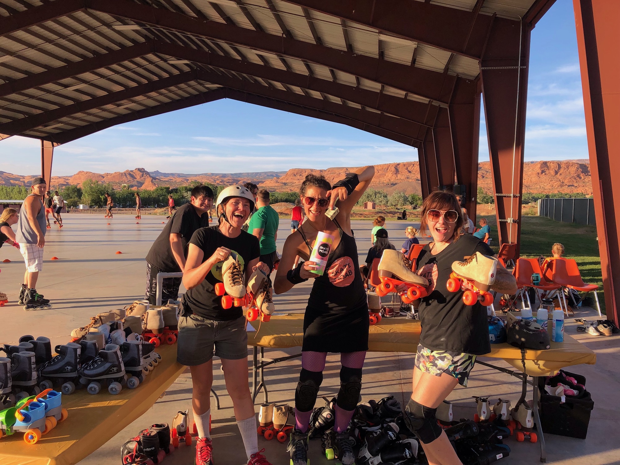 Three organizers are pictured at the skate rental and cleaning table during a skating night; they're each doing a goofy pose and smiling.