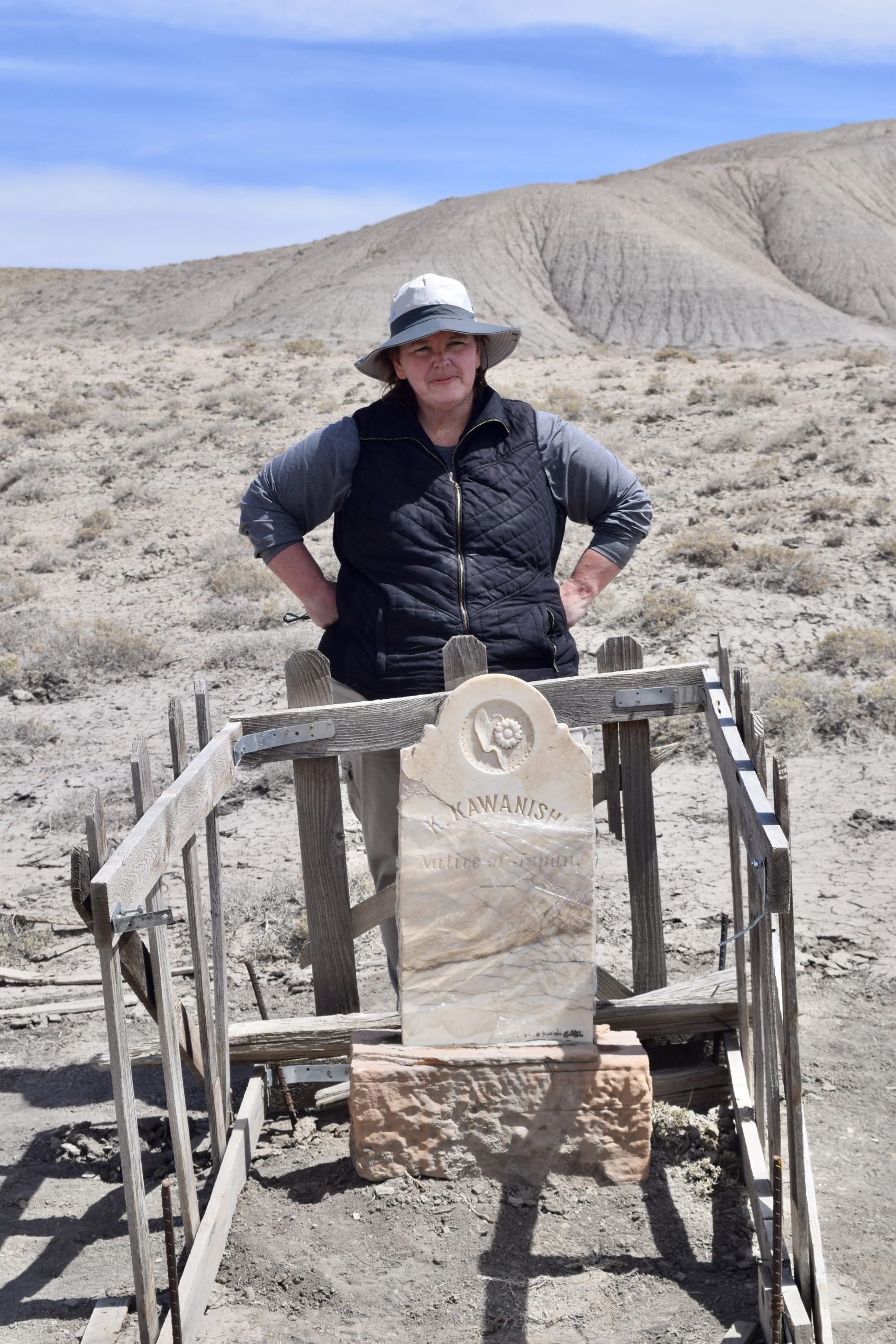 Amy Barry is wearing a bucket hat and vest. She stands behind the fixed headstone with her hands on her hips and a small smile on her face.