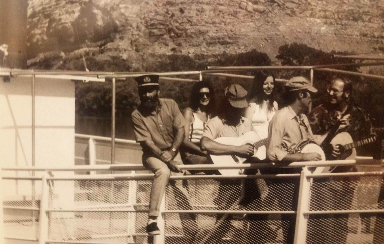 A group of six people are pictured on an old tour boat. Three of them are playing guitars. Tex McClatchy, identifiable because of his beard, wears a captains hat as he leans over the side of the boat.