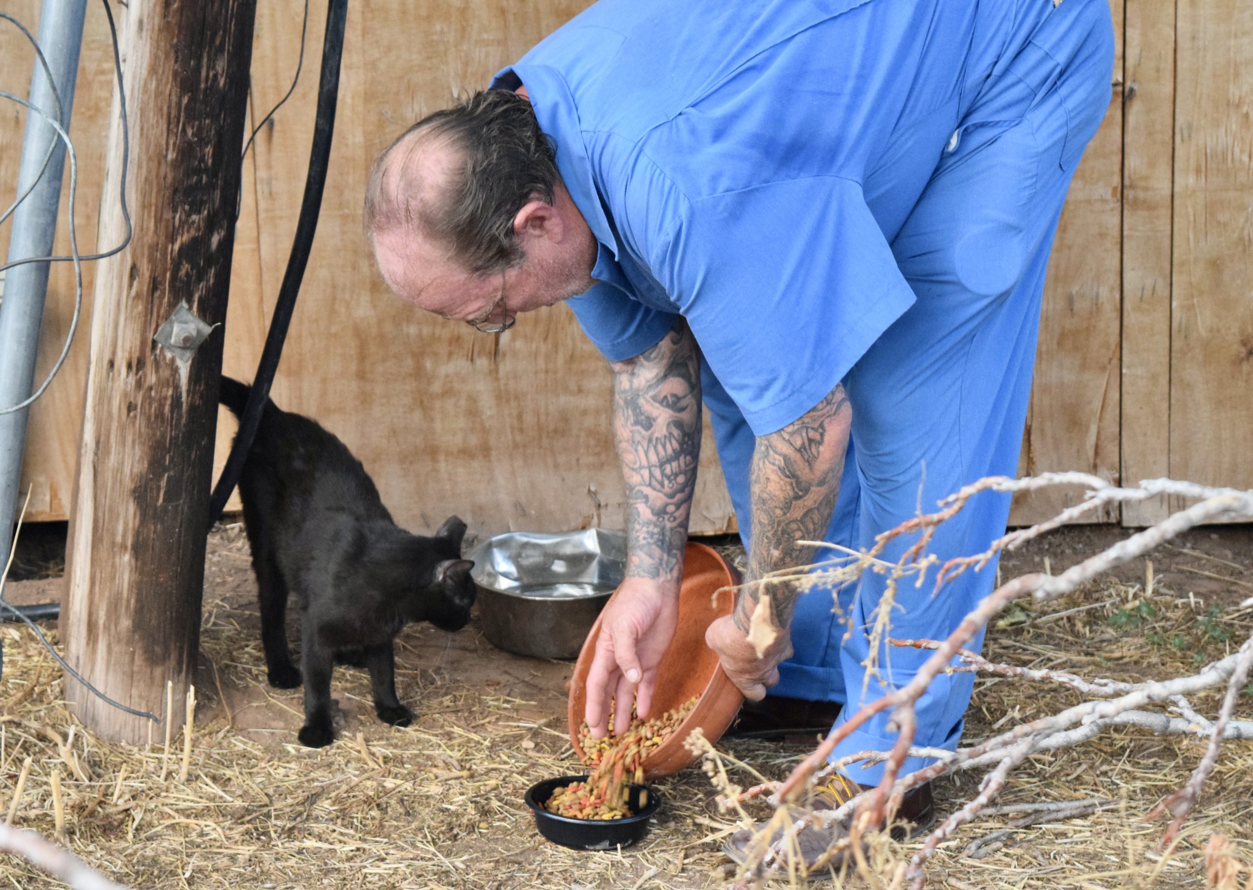 Travis Garcia, dressed in blue scrubs, pours cat food into a small dish. A black cat waits patiently nearby.