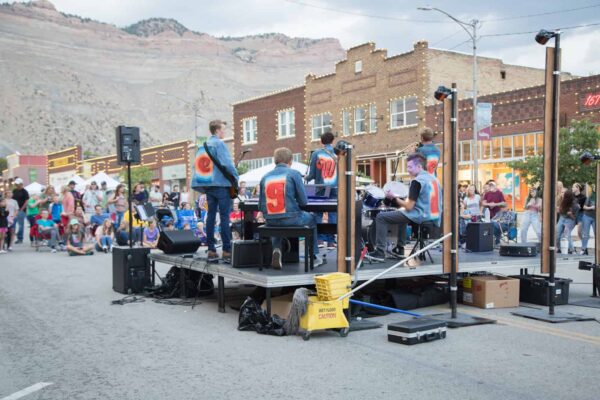 A band of four people play on a small stage in front of a crown on Helper's Main St: there are historic buildings in the background.