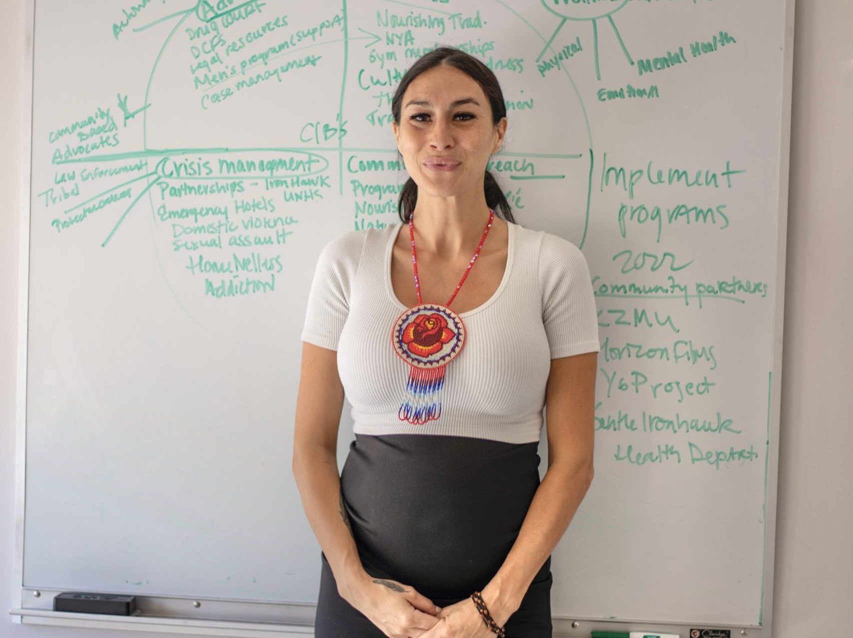 Kristen, dressed in a white t-shirt and pencil skirt, stands in front of a whiteboard filled with ideas for the nonprofit: words like "advocacy" surround her.