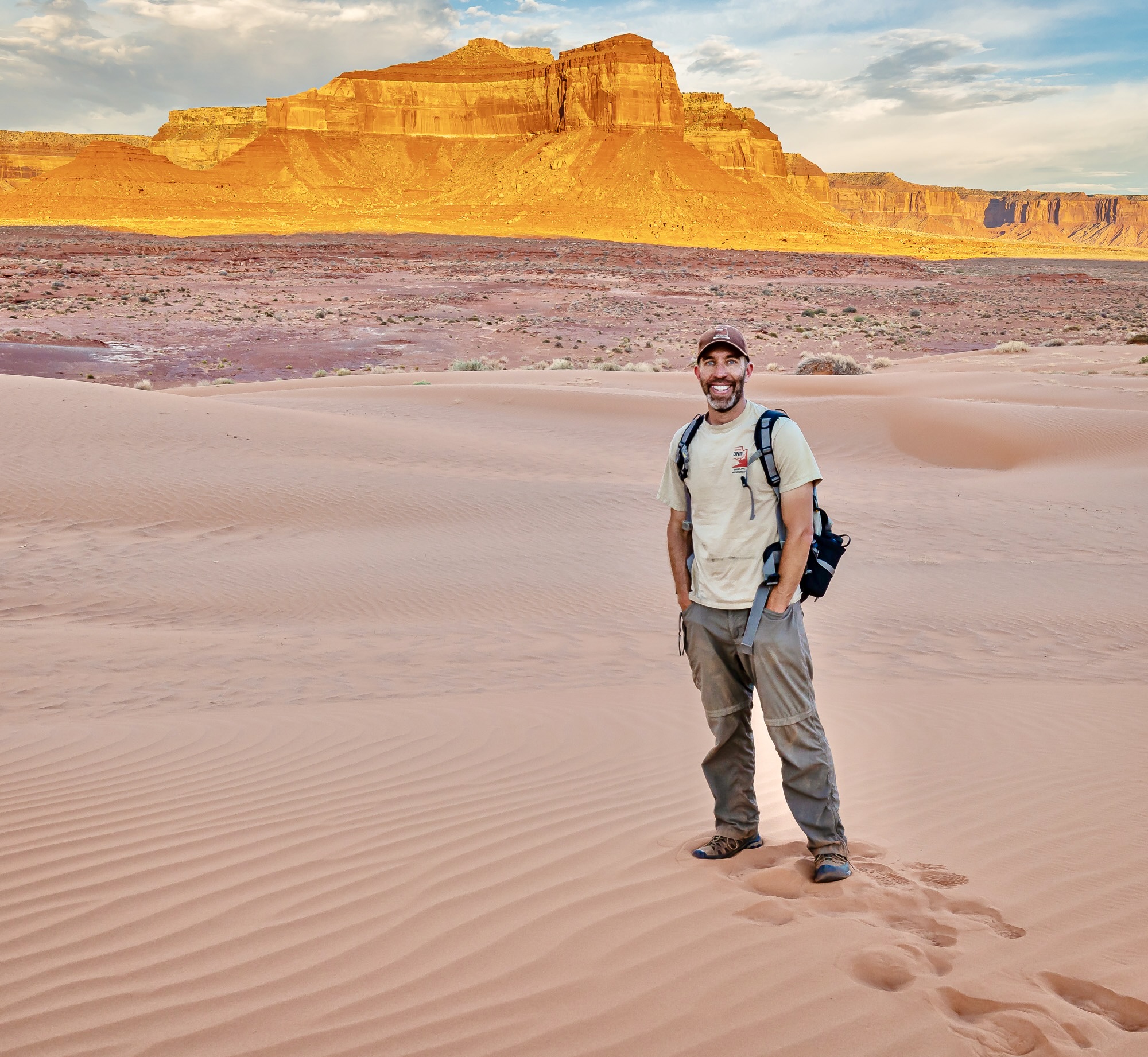 Scott Gibson stands in the sand in front of an impressive red rock spire: you can see his footprints through the sand.