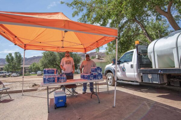 Two men stand underneath an orange tent; they have an array of water bottles on a table.