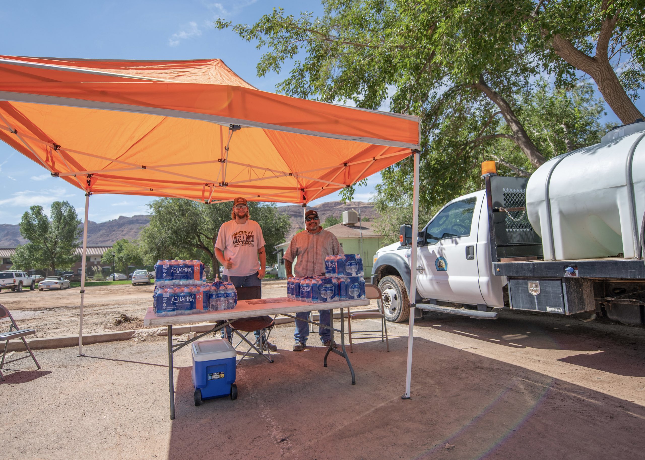 Two men stand underneath an orange tent; they have an array of water bottles on a table.