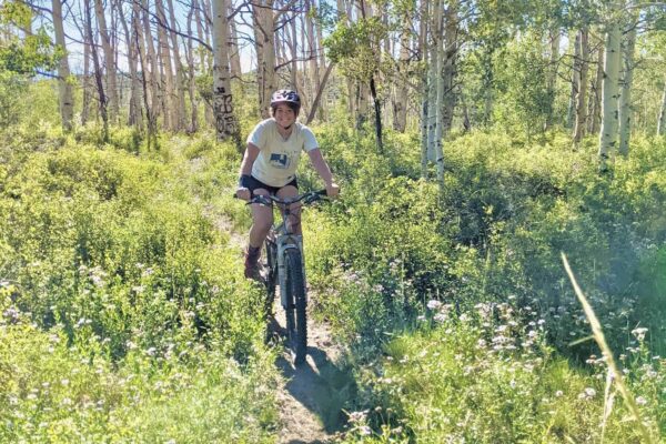 A mountain biker bikes through a field of flowers and aspen trees. They're smiling.