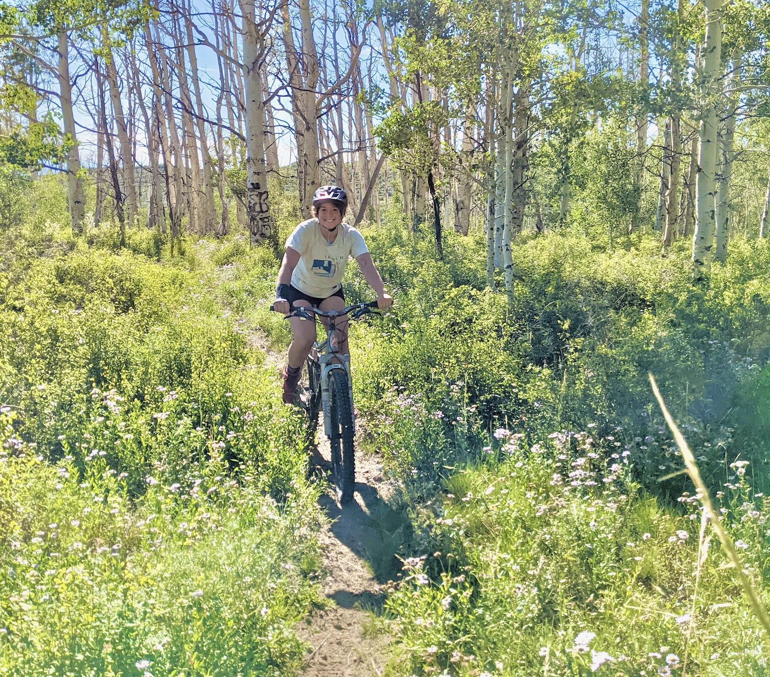 A mountain biker bikes through a field of flowers and aspen trees. They're smiling.