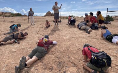 Steve Leavitt is standing in the middle of a circle of people who are all laid out on the ground, observing lichen on slickrock.