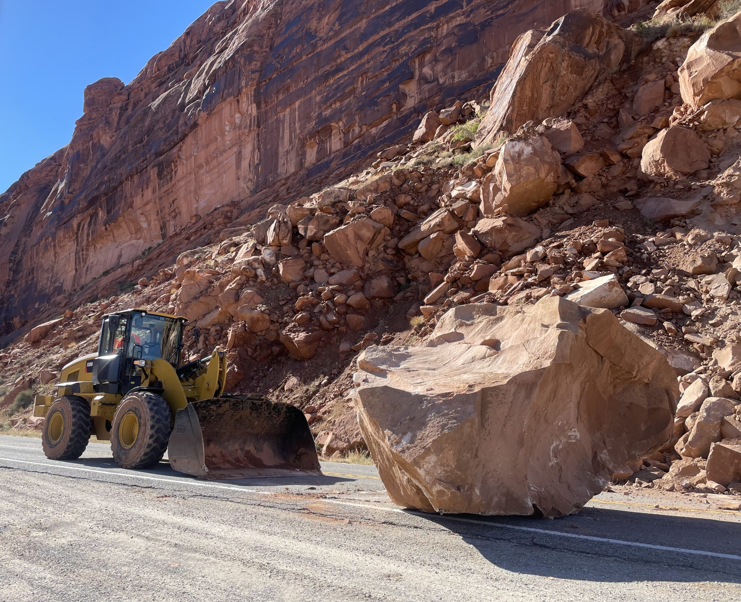 A large boulder—larger than the excavator pictured next to it—on the road.