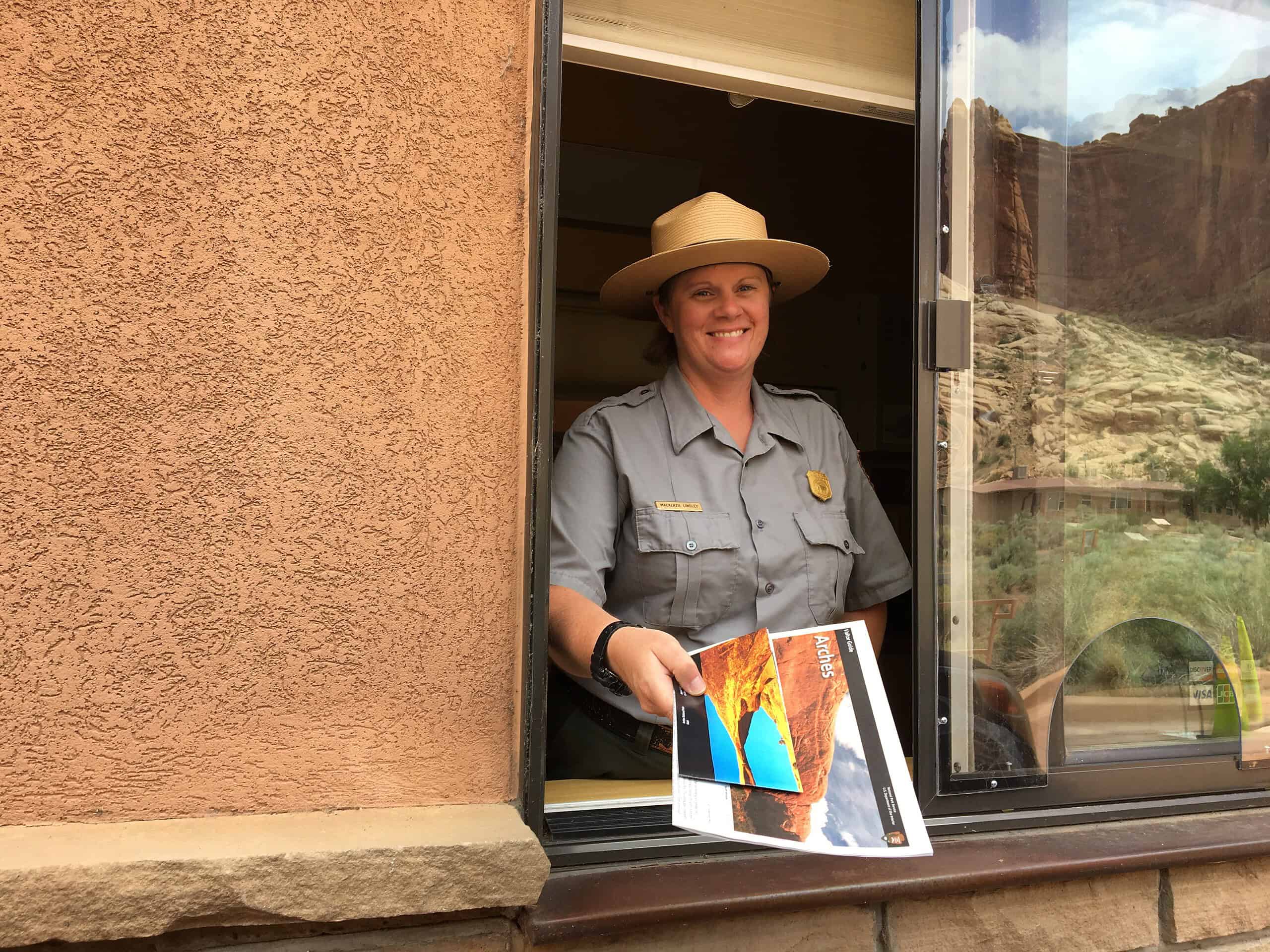 Park ranger in uniform smiling at a window, holding a brochure with a scenic view in the background.