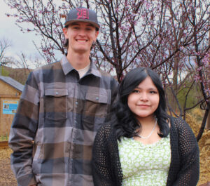 Two people smiling outdoors with blossoming trees in the background, one wearing a cap, standing on a wood-chip path.