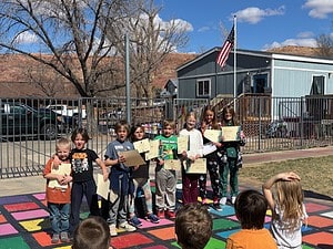 Children holding certificates outdoors at a school event, standing on colorful tiles under a clear blue sky.