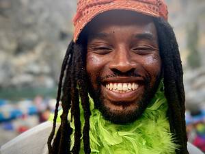 Smiling person with dreadlocks and colorful decorations, wearing a feathered scarf and cap outdoors.