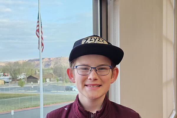 Smiling person in a cap by a window with an American flag and scenic view outside.