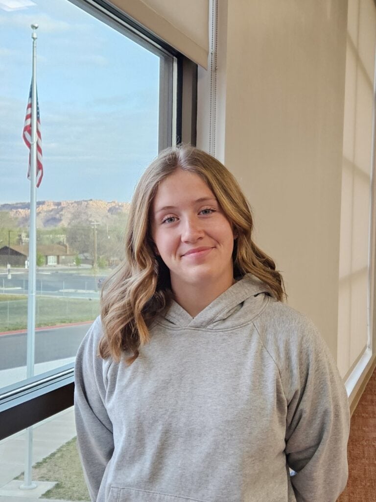Ellie Cornelius in a gray hoodie beside a window with an American flag visible outside.