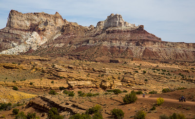 Rugged desert landscape with layered rock formations and distant ATV riders under a clear sky.