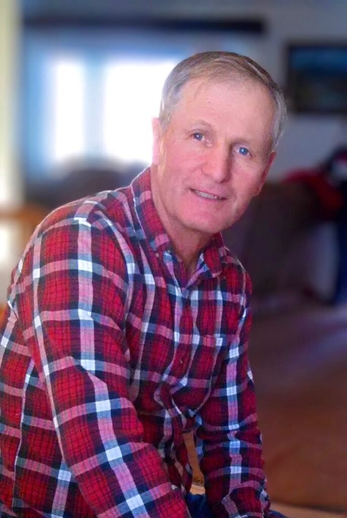 Smiling man in a red plaid shirt seated indoors with soft lighting in the background.
