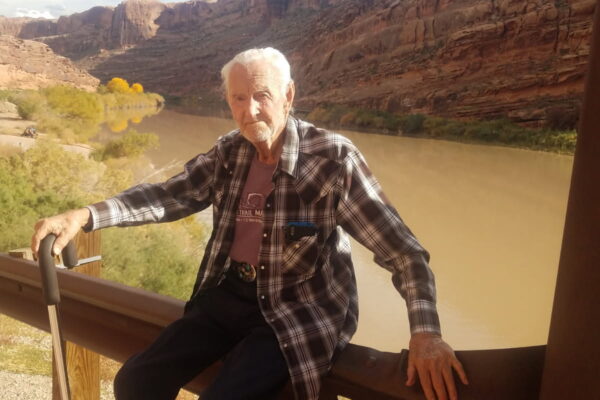 Elderly man with cane sits by a river, surrounded by scenic rocky landscape under a blue sky.