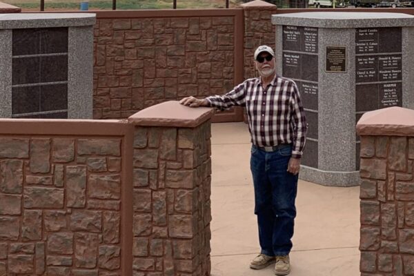Man standing by brick wall with mountainous backdrop, cloudy skies, and rural landscape.