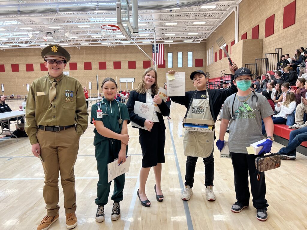 Students in costume at a school event, dressed as various professionals, stand proudly with certificates.