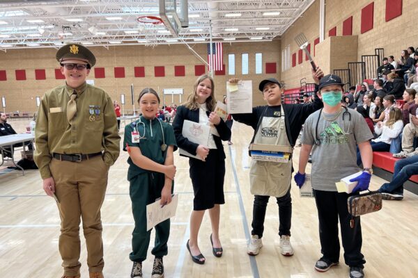 Students in costume at a school event, dressed as various professionals, stand proudly with certificates.
