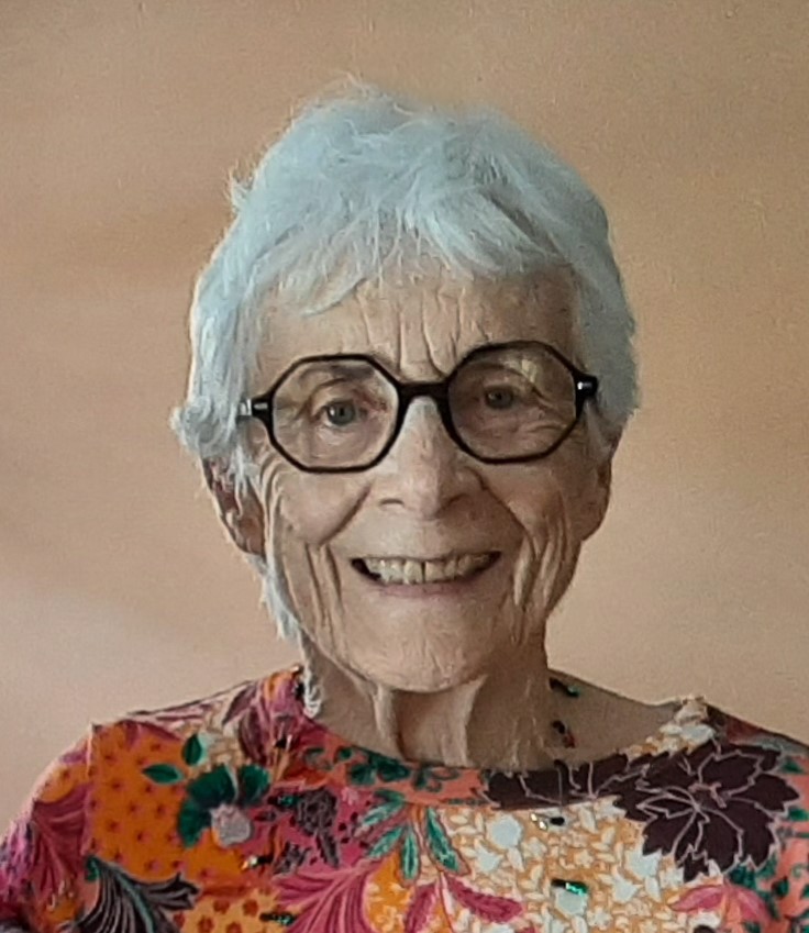 Elderly woman with white hair, glasses, and a vibrant floral top smiles warmly against a neutral background.