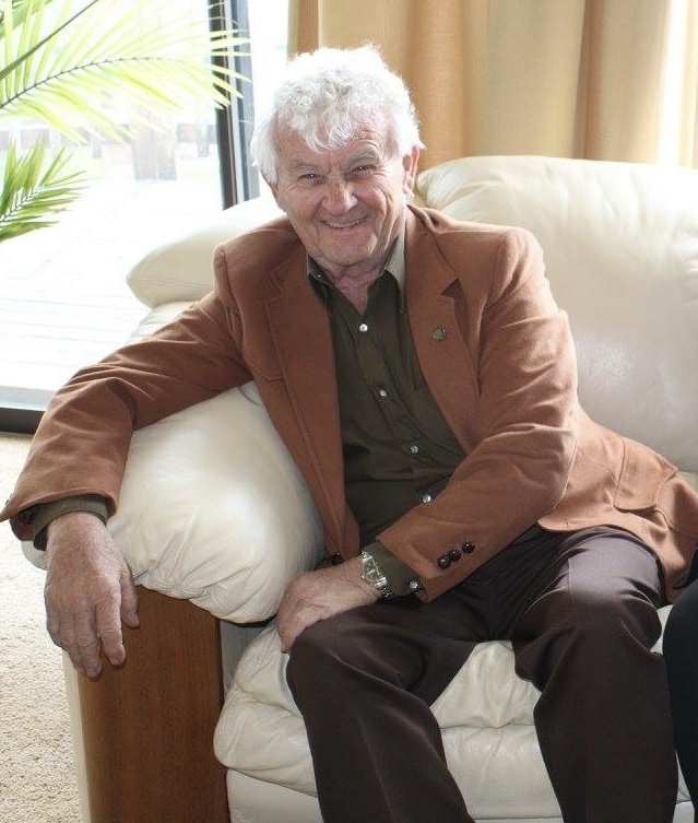 Elderly man in brown blazer sitting on sofa, smiling warmly at camera, indoor setting with natural light.