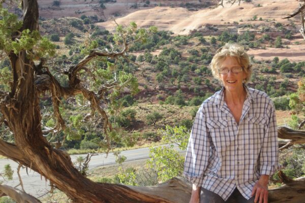 Smiling person in plaid shirt sitting by tree with scenic desert landscape in background.