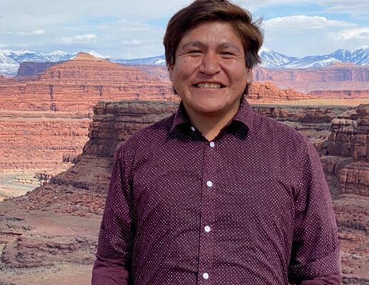 Smiling person in a purple shirt standing in front of a stunning canyon landscape with clear skies.