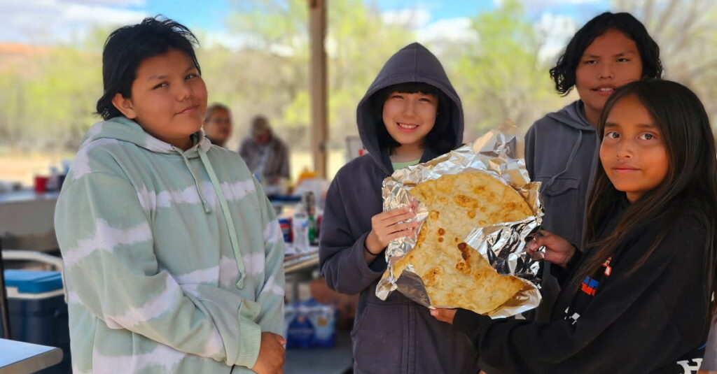 Group of kids proudly holding a large tortilla at an outdoor gathering under a metal-roofed shelter.
