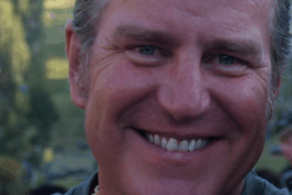 Smiling man outdoors with blurred green landscape in background, wearing a dark shirt, enjoying sunny day.