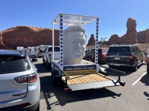 Large head sculpture on trailer with Make America Wait Again sign, parked near red rock formations under clear blue sky.