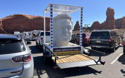 Large head sculpture on trailer with Make America Wait Again sign, parked near red rock formations under clear blue sky.