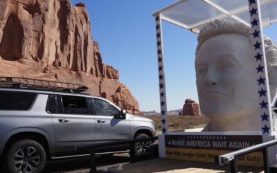 SUV beside large statue and rock formations under blue sky in desert landscape.