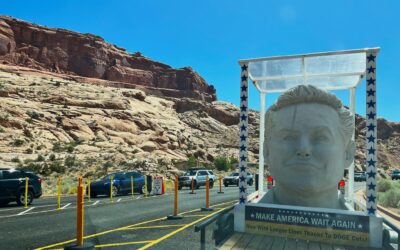 Large sculpture on trailer with Make America Wait Again sign, desert landscape, cars lined up. Clear blue sky.