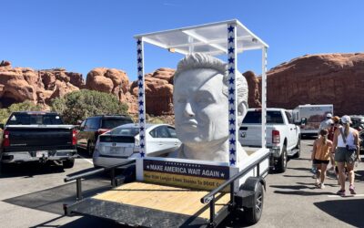 Bust of a man on trailer reading Make America Wait Again, in a parking lot with red rock formations and people nearby.