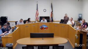 Meeting in a council chamber with people discussing at a curved table, flags and clock in background.