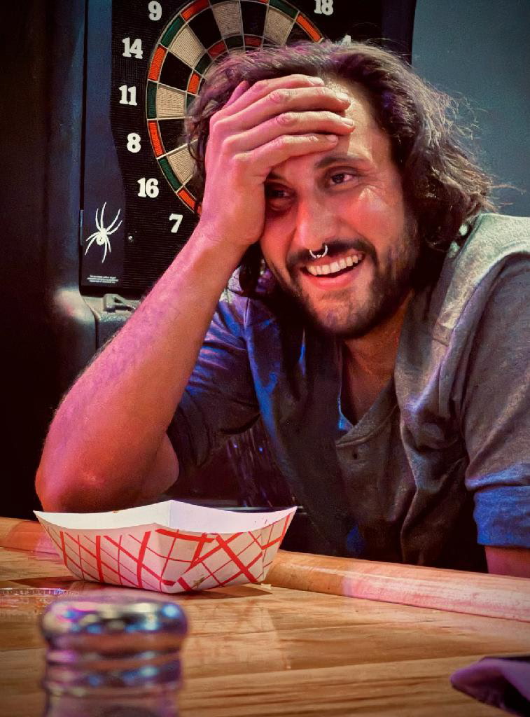 Man smiling at a bar with a dartboard in the background, holding his head playfully.