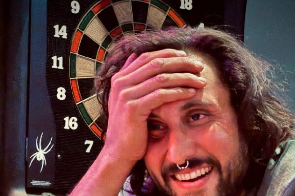 Man smiling at a bar with a dartboard in the background, hand on head, and paper food tray on the counter.