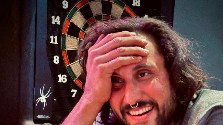 Man smiling at a bar with a dartboard in the background, hand on head, and paper food tray on the counter.