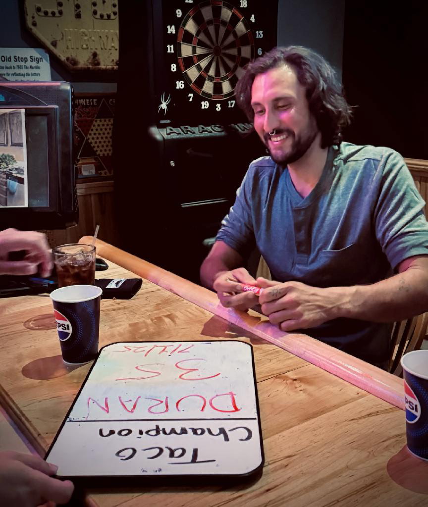 Man smiling at a table with a dartboard in the background, next to a whiteboard and drinks.