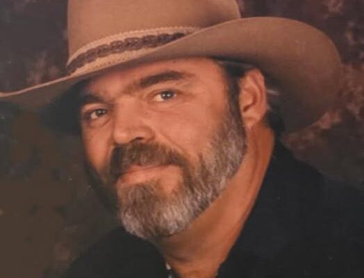 Man in cowboy hat with beard, wearing a dark shirt, posing against a neutral background.